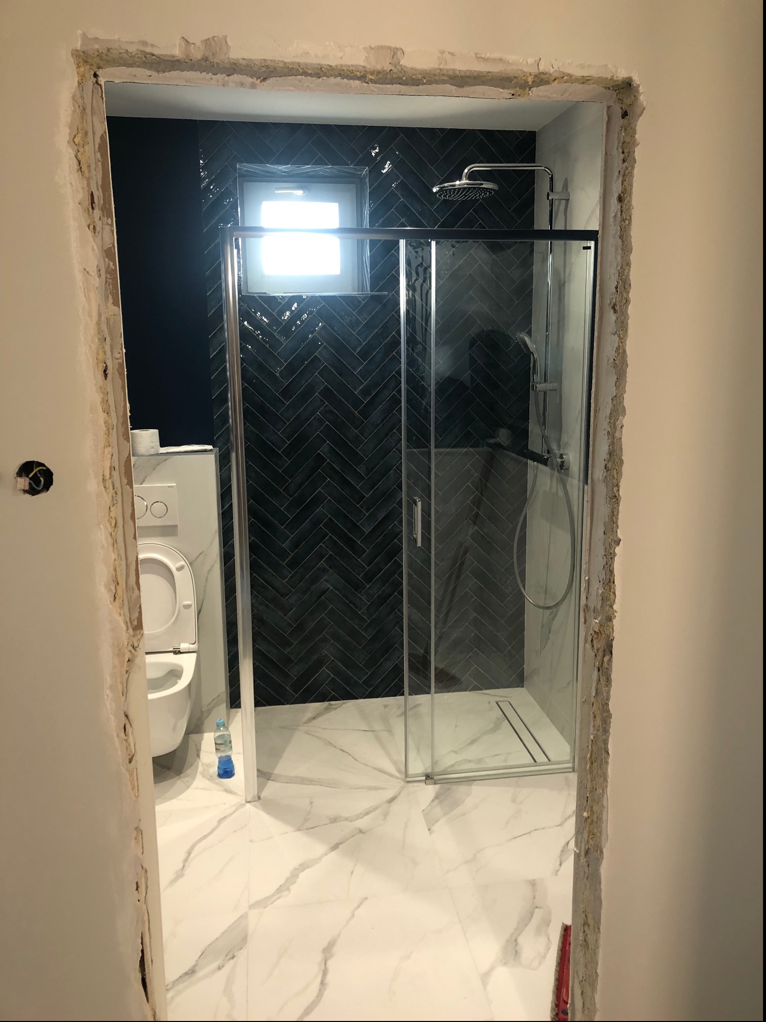 Łazienka w trakcie remontu: widok przez surową futrynę na kabinę prysznicową z czarnymi płytkami ułożonymi w jodełkę na ścianie i białą marmurową podłogą. Uwagę zwraca butelka wody mineralnej...