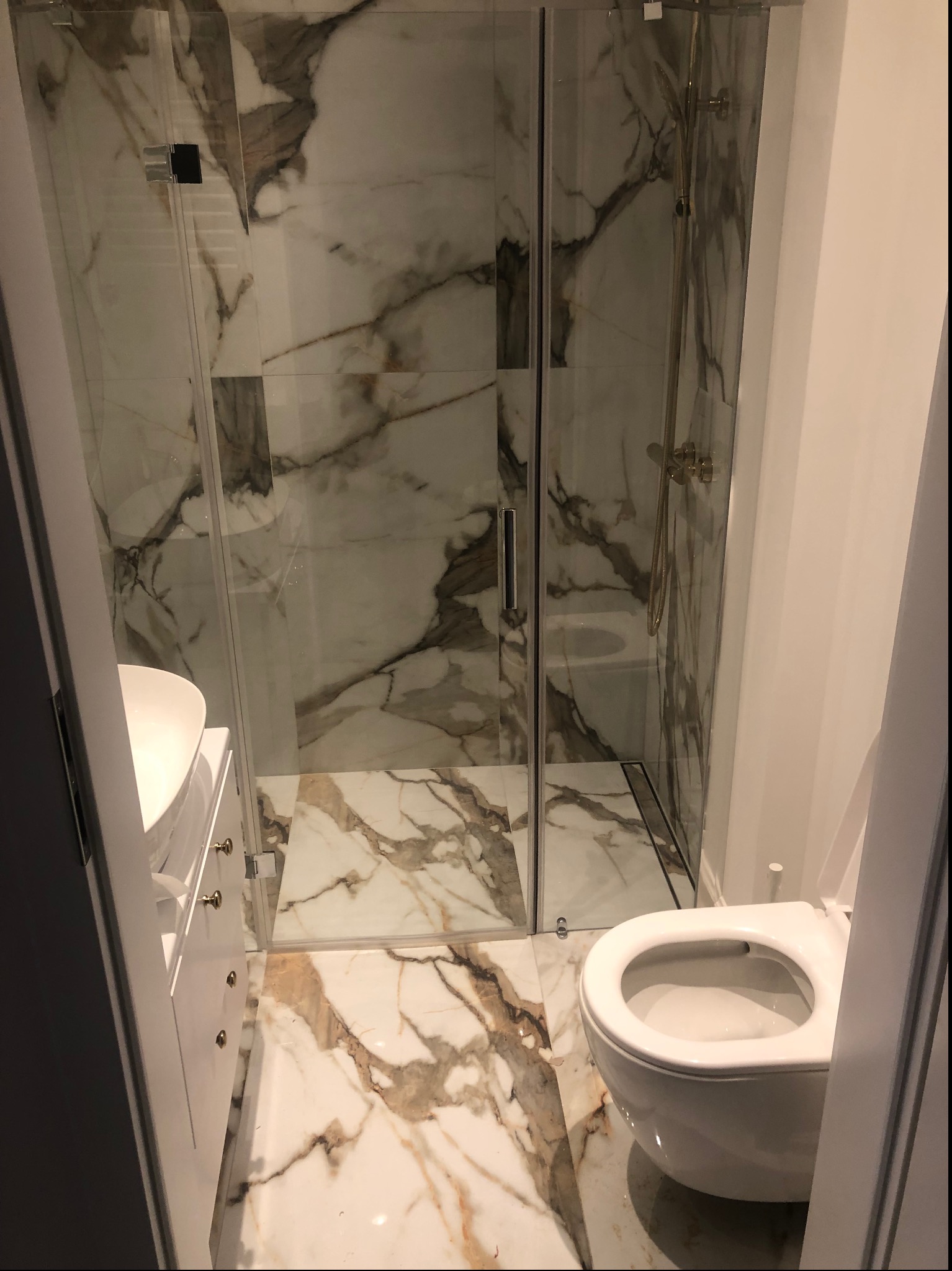 Wąska łazienka z kabiną prysznicową ze złotą armaturą, umywalką z szafką i podwieszaną toaletą, wykończona płytkami imitującymi marmur Calacatta.