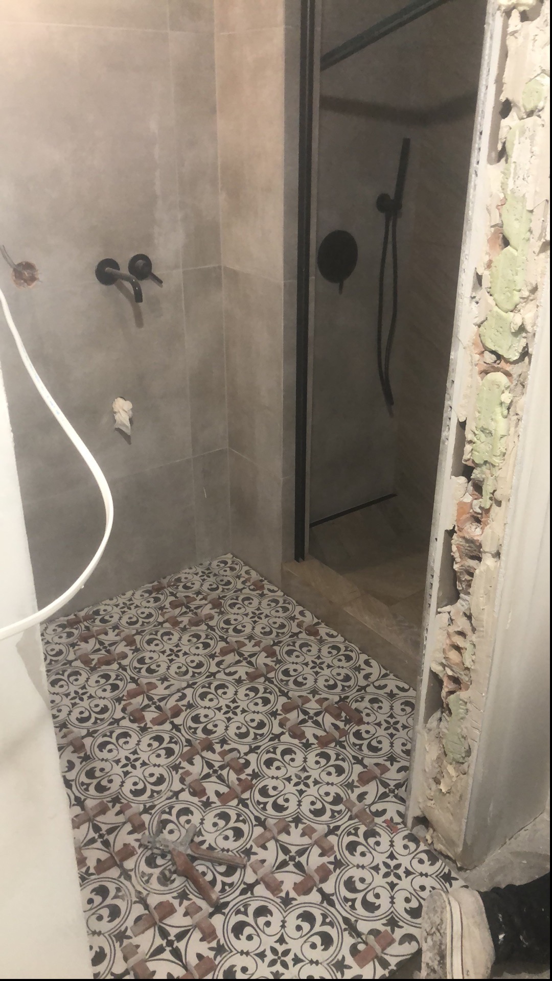 Wnętrze łazienki w trakcie remontu: położona dekoracyjna podłoga z wzorem, widoczne czarne armatura, kabina prysznicowa z czarną ramą oraz fragment ściany z uszkodzonym tynkiem.