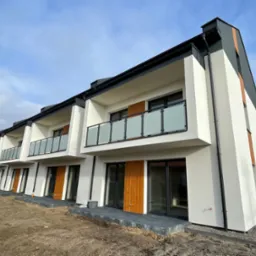 Nowoczesne szeregowe domy z białymi elewacjami, szarymi elementami dachu i drewnianymi akcentami, balkony z mlecznymi balustradami, teren wokół w trakcie zagospodarowywania.