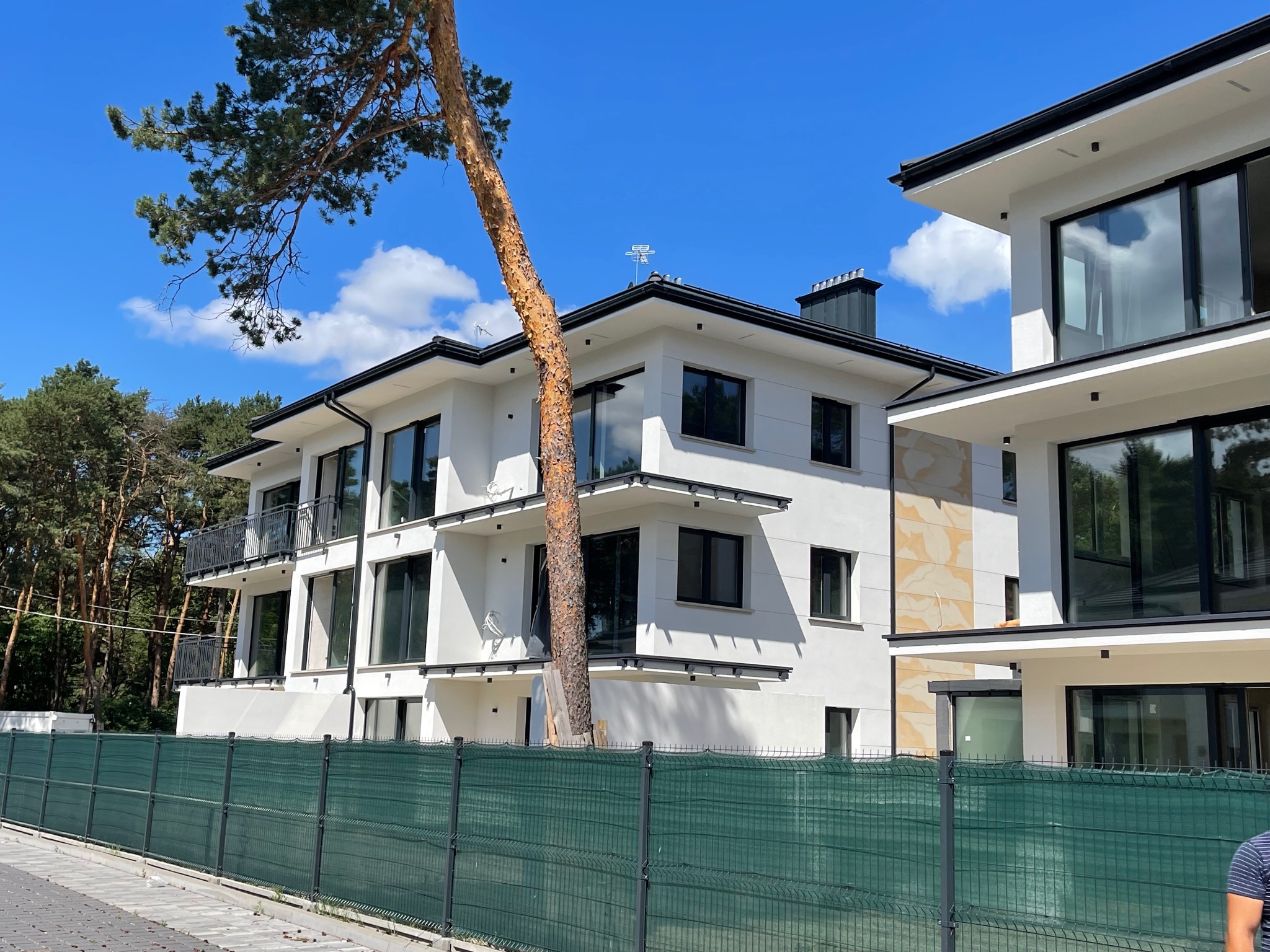 Nowoczesny, dwupiętrowy budynek z białą elewacją, czarnymi ramami okiennymi i balkonami, widziany zza zielonego ogrodzenia. Obok budynku rośnie wysokie drzewo, a w tle widać las pod błękitnym niebem.