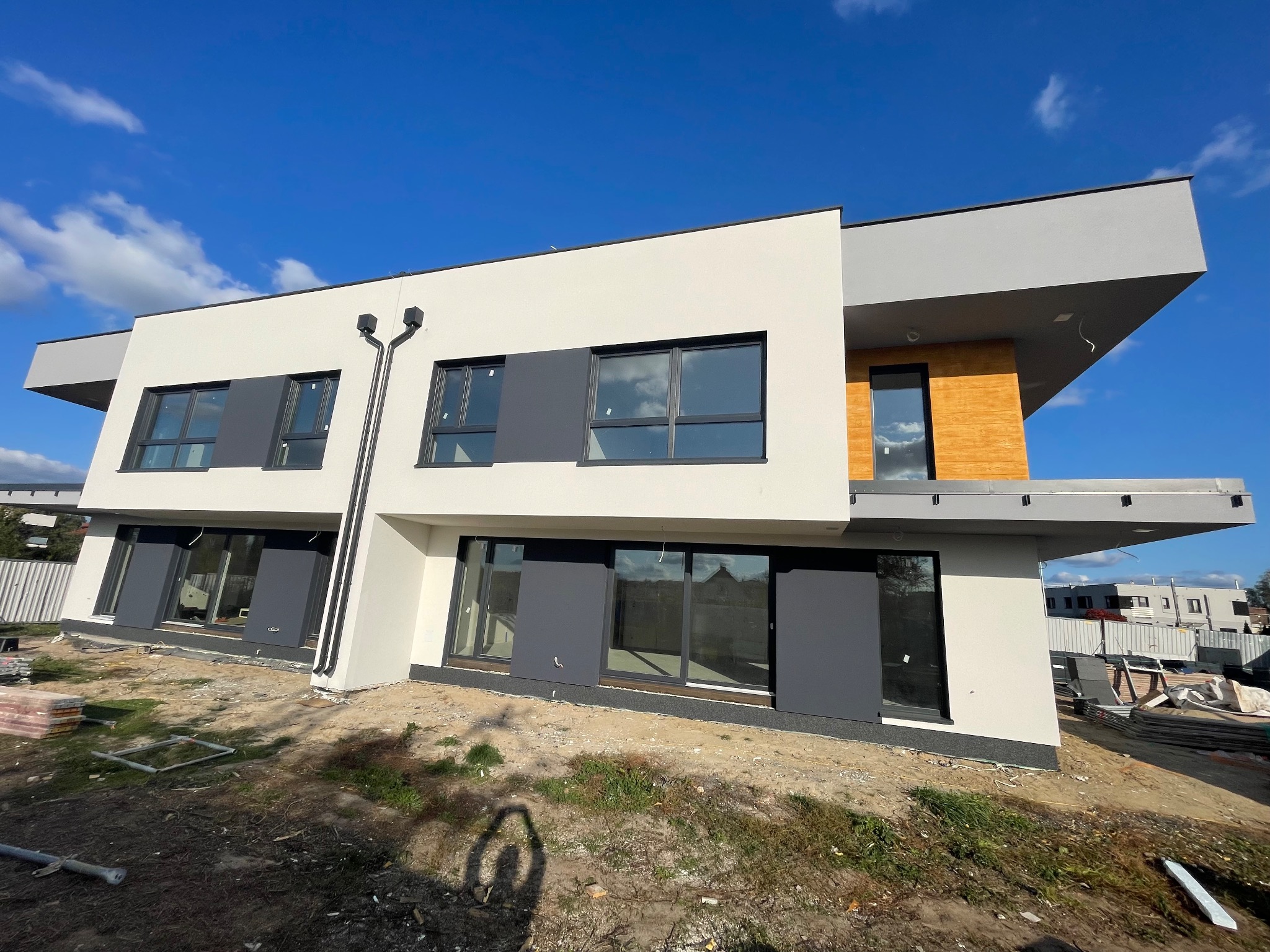 Nowoczesny, dwukondygnacyjny dom z płaskim dachem, białą elewacją, szarymi akcentami i dużymi oknami, w trakcie prac wykończeniowych.