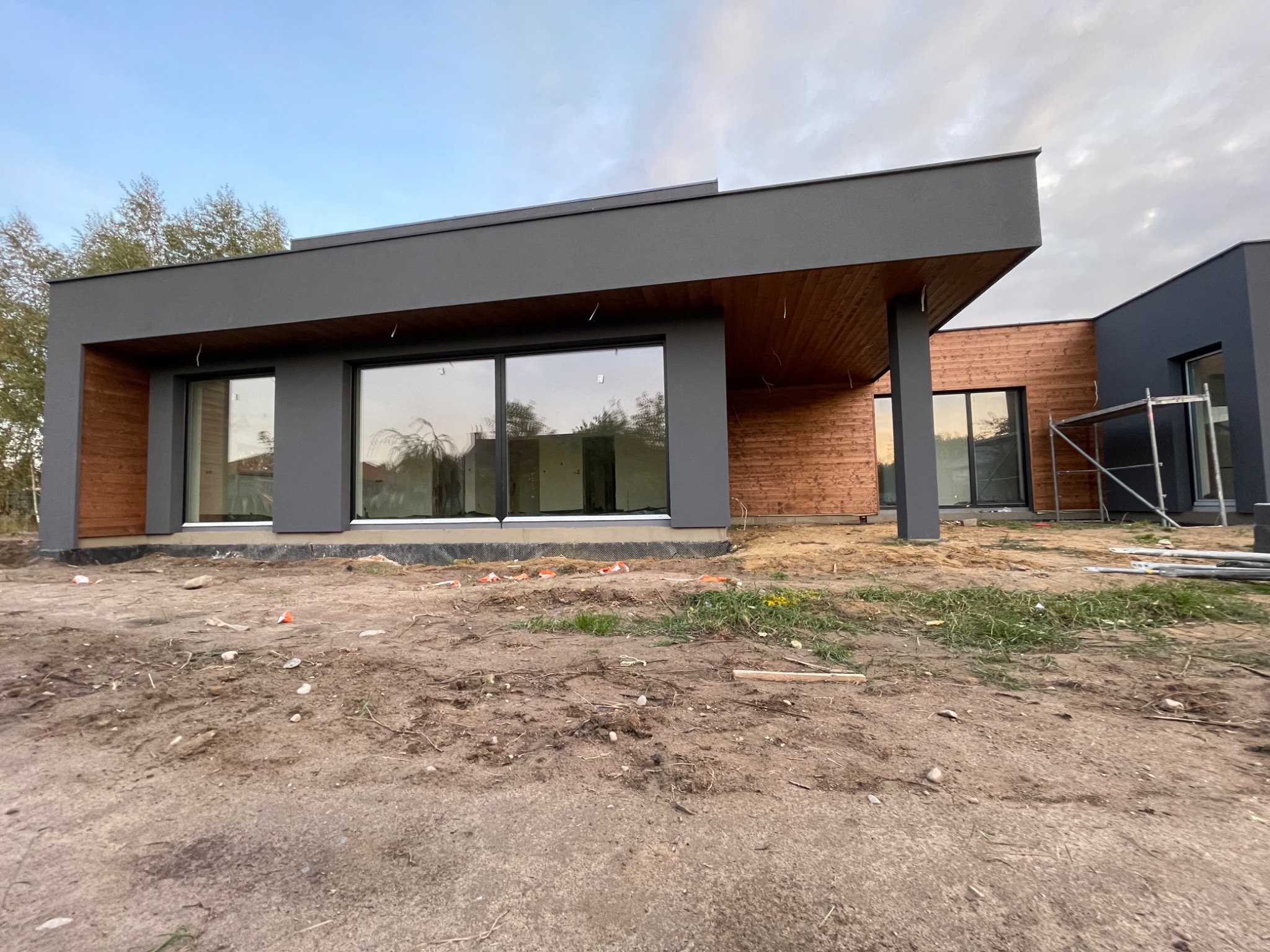 Nowoczesny dom jednorodzinny z szarą elewacją i drewnianym wykończeniem fragmentów ścian, widoczne okna z odbiciem nieba, teren wokół domu w trakcie prac wykończeniowych, ziemia i resztki...