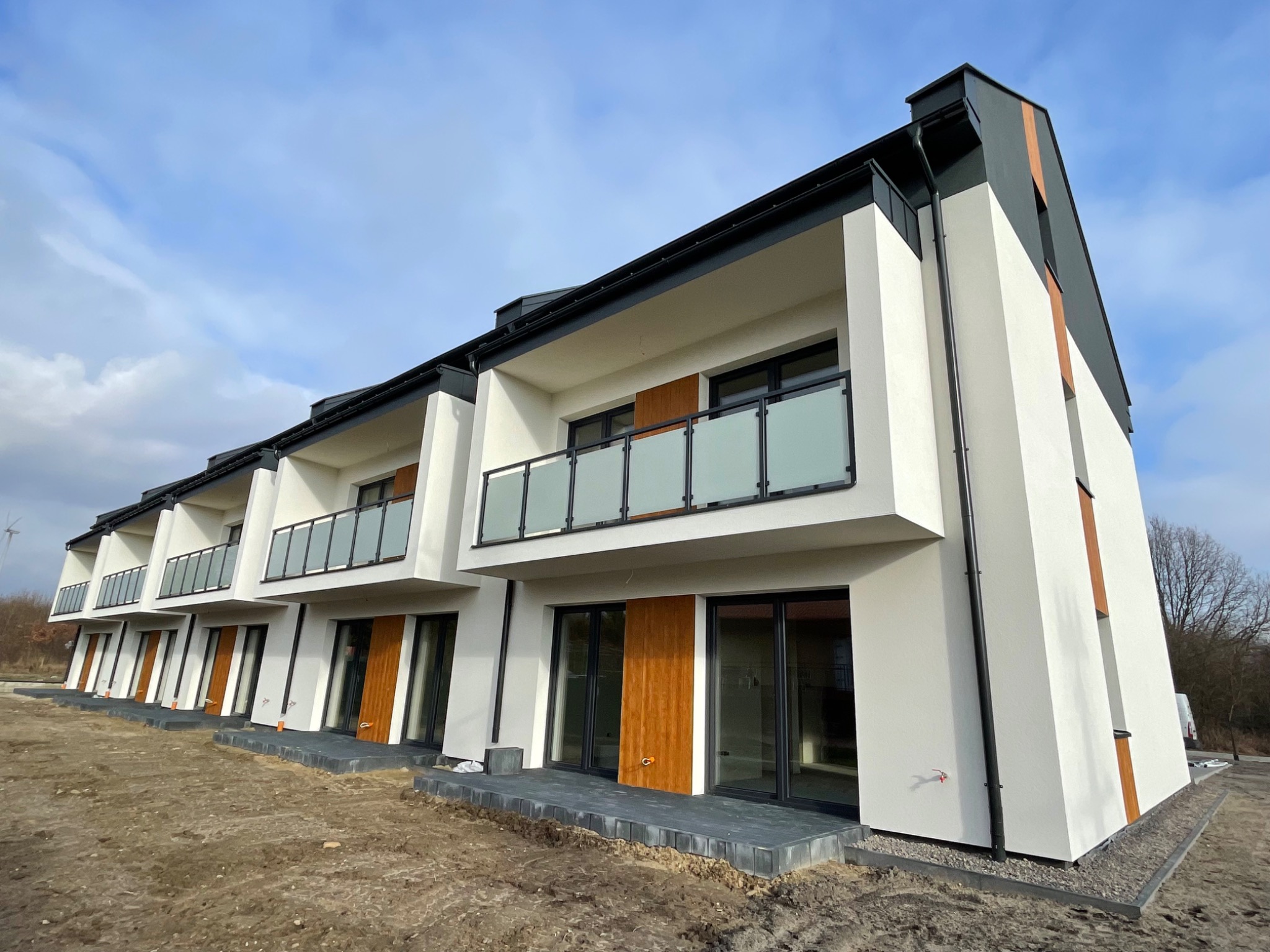 Nowoczesne szeregowe domy z białymi elewacjami, szarymi elementami dachu i drewnianymi akcentami, balkony z mlecznymi balustradami, teren wokół w trakcie zagospodarowywania.