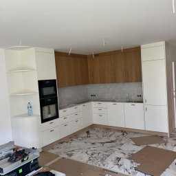 Visual trend meble na wymiar - Wnętrze kuchni w trakcie montażu: białe i drewniane szafki, marmurowe płytki podłogowe zabezpieczone kartonem, widoczne narzędzia i akcesoria montażowe, niedokończone wykończenie sufitu.