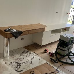 Visual trend meble na wymiar - Montaż drewnianego blatu z szufladami na chromowanej nodze i białej szafki; widoczny odkurzacz przemysłowy i narzędzia na podłodze zabezpieczonej kartonem.