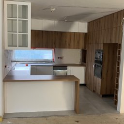 Visual trend meble na wymiar - Wnętrze kuchni w trakcie montażu, widoczne drewniane i białe szafki, wyspa kuchenna z blatem, sprzęt AGD w folii ochronnej, surowa podłoga przed wykończeniem.