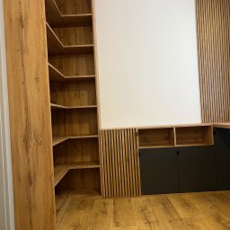 Visual trend meble na wymiar - Zabudowa ściany z drewnianymi lamelami i szafkami z czarnymi frontami oraz regał narożny z półkami z płyty drewnopodobnej.