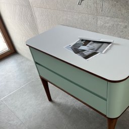 Visual trend meble na wymiar - Pastelowa szafka łazienkowa z dwoma szufladami na drewnianych nóżkach, na blacie leżą zdjęcia aranżacji wnętrz, w tle płytki imitujące kamień.