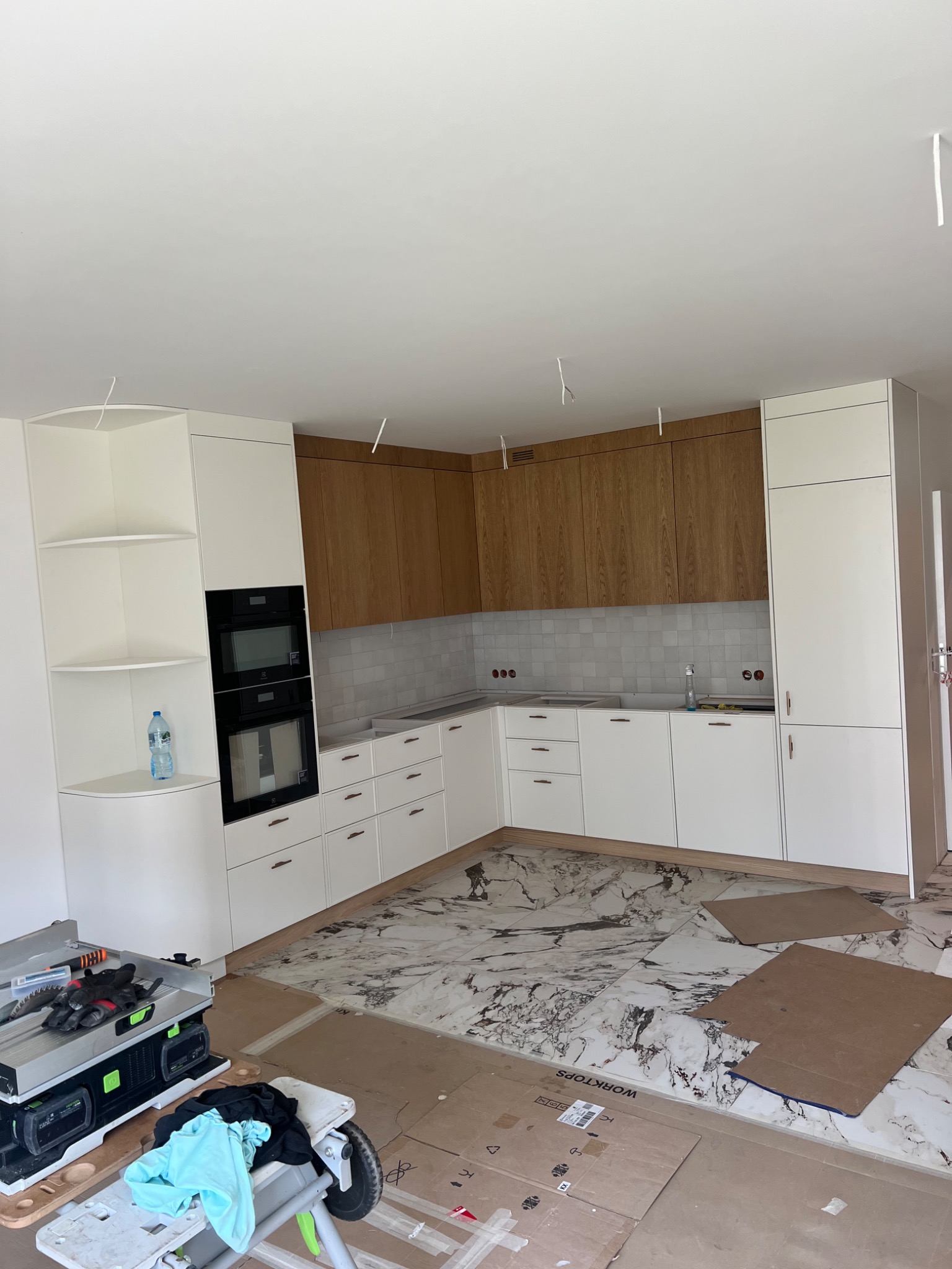 Wnętrze kuchni w trakcie montażu: białe i drewniane szafki, marmurowe płytki podłogowe zabezpieczone kartonem, widoczne narzędzia i akcesoria montażowe, niedokończone wykończenie sufitu.