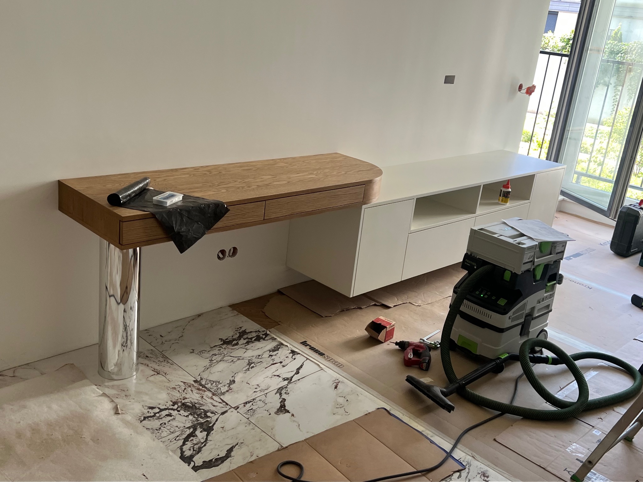 Montaż drewnianego blatu z szufladami na chromowanej nodze i białej szafki; widoczny odkurzacz przemysłowy i narzędzia na podłodze zabezpieczonej kartonem.