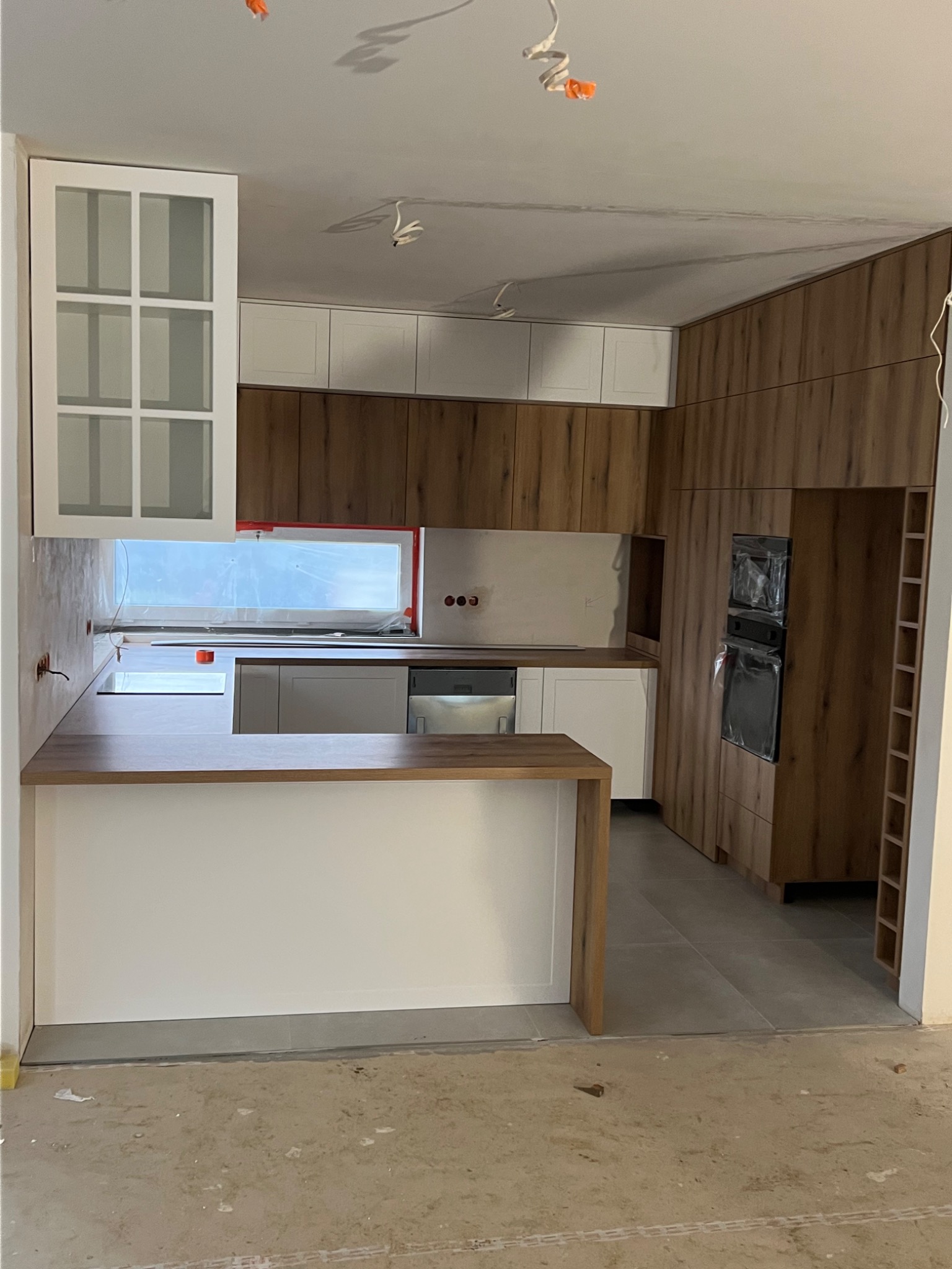 Wnętrze kuchni w trakcie montażu, widoczne drewniane i białe szafki, wyspa kuchenna z blatem, sprzęt AGD w folii ochronnej, surowa podłoga przed wykończeniem.