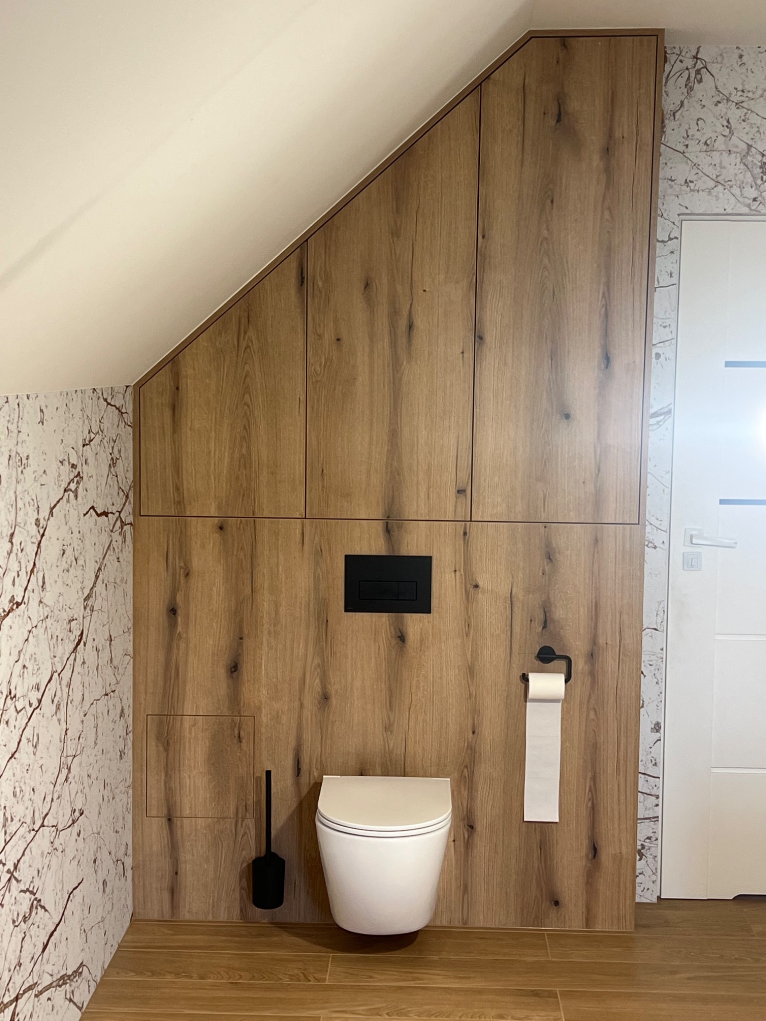 Nowoczesna łazienka na poddaszu z zabudową z drewnopodobnych paneli, ukrywającą stelaż WC i z szafkami pod skosem, z widoczną toaletą, uchwytem na papier toaletowy i szczotką do WC w kolorze czarnym.