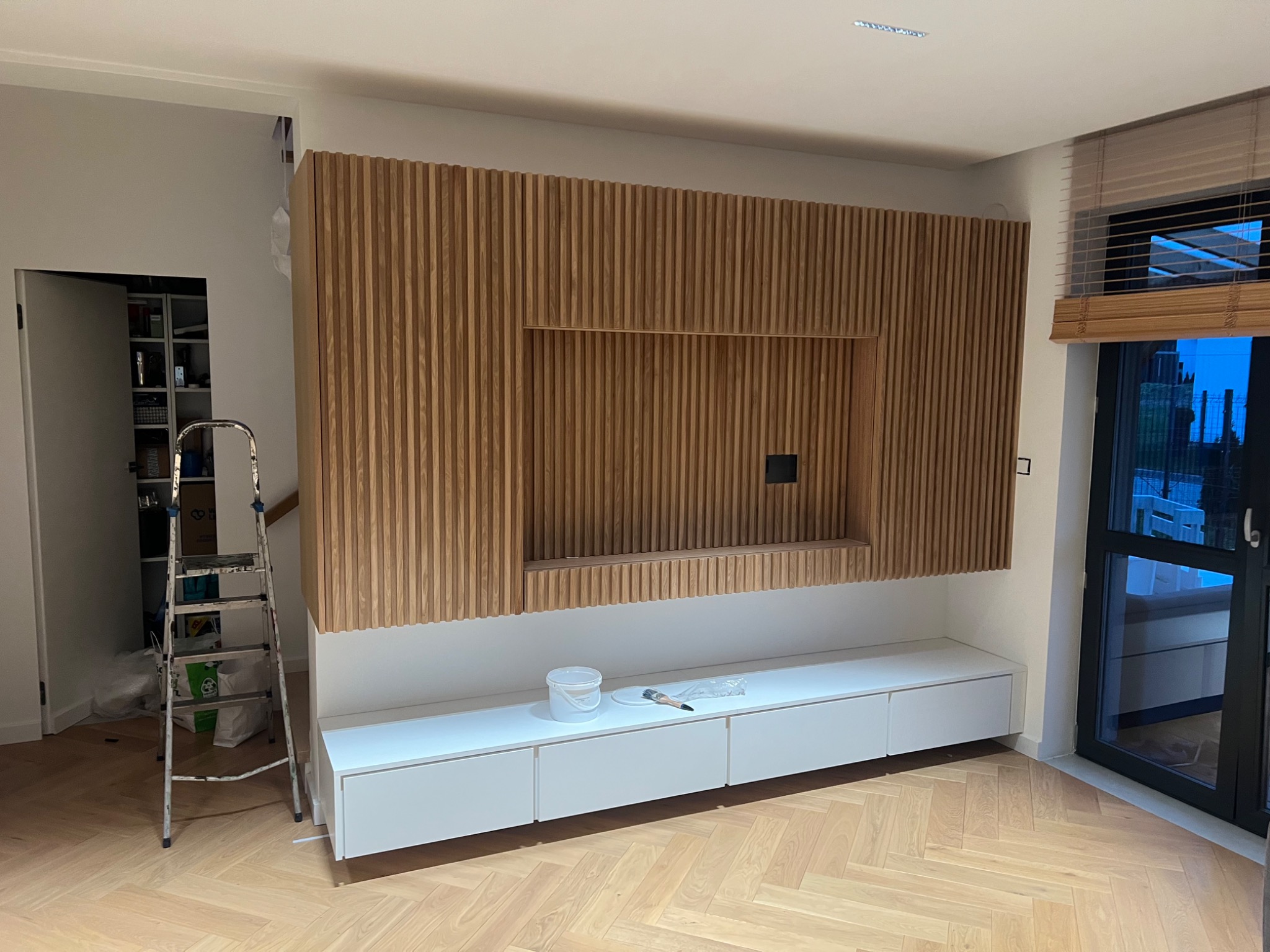 Ściana z drewnianych lameli z wnęką na telewizor, biała szafka RTV, drabina malarska i wiadro z farbą w pomieszczeniu z parkietem ułożonym w jodełkę.