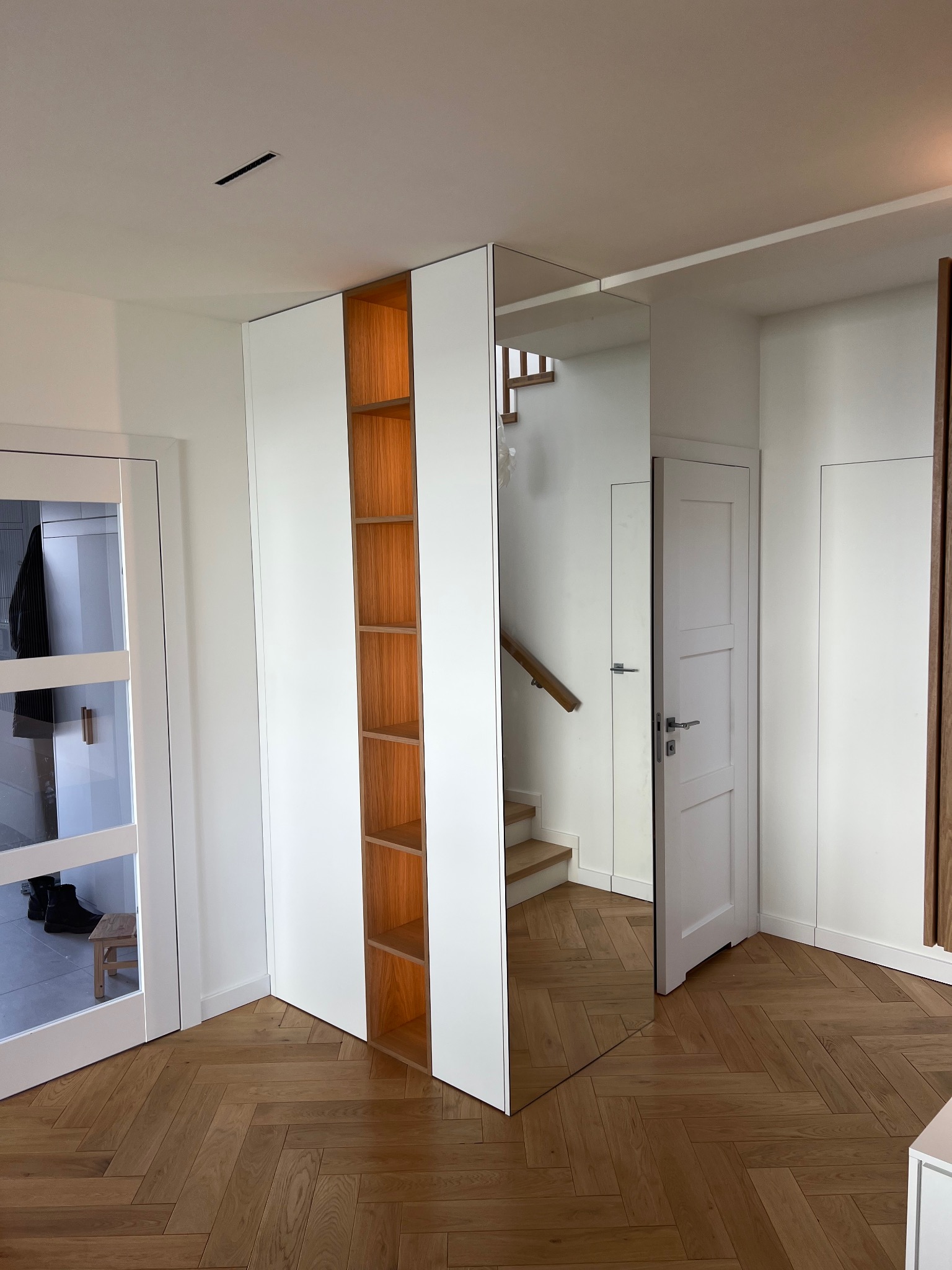 Nowoczesna zabudowa przedpokoju z szafą z białymi frontami i otwartymi półkami z drewna, lustrzane drzwi odbijają schody, parkiet ułożony we wzór jodełki.