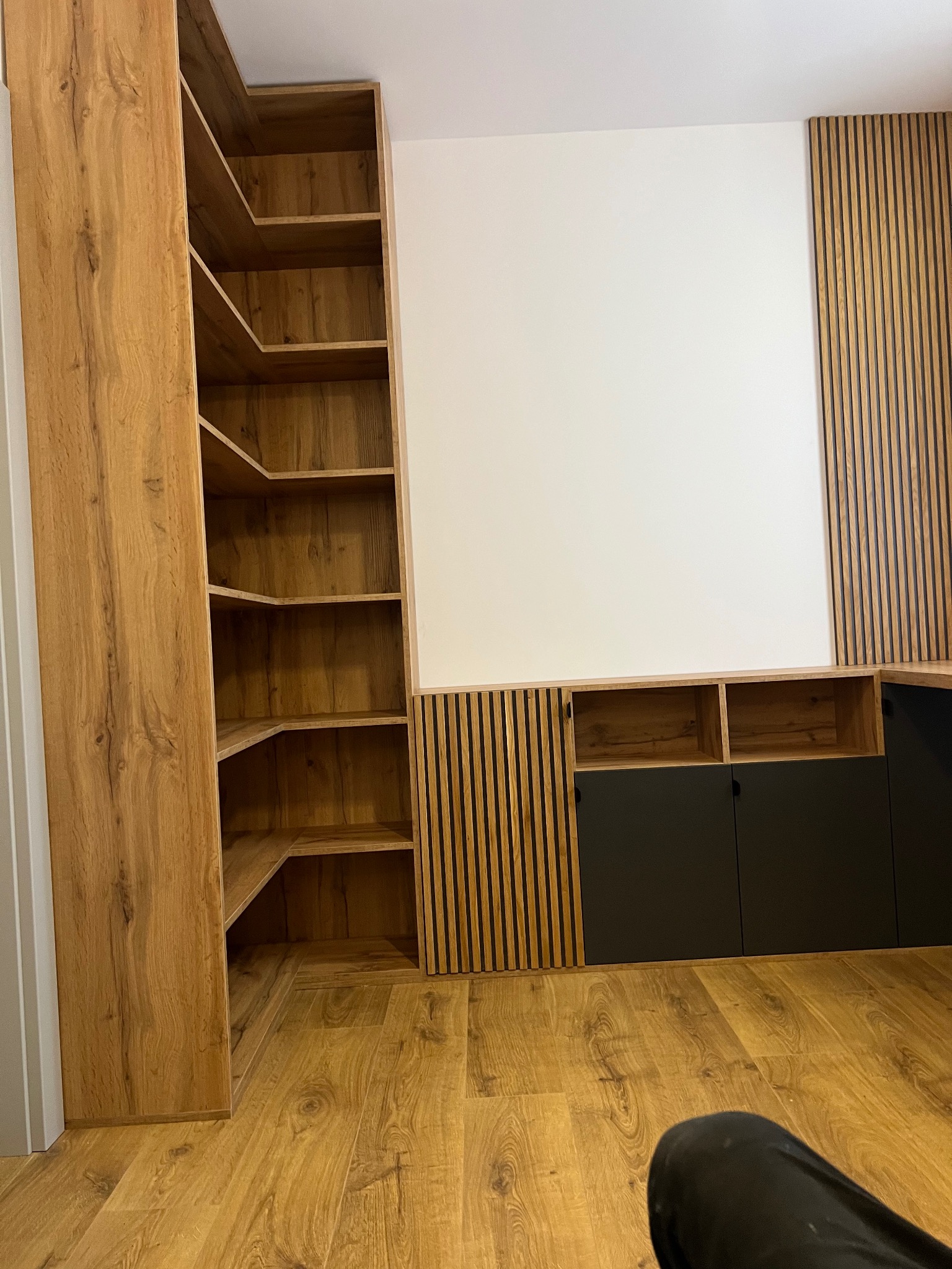 Zabudowa ściany z drewnianymi lamelami i szafkami z czarnymi frontami oraz regał narożny z półkami z płyty drewnopodobnej.