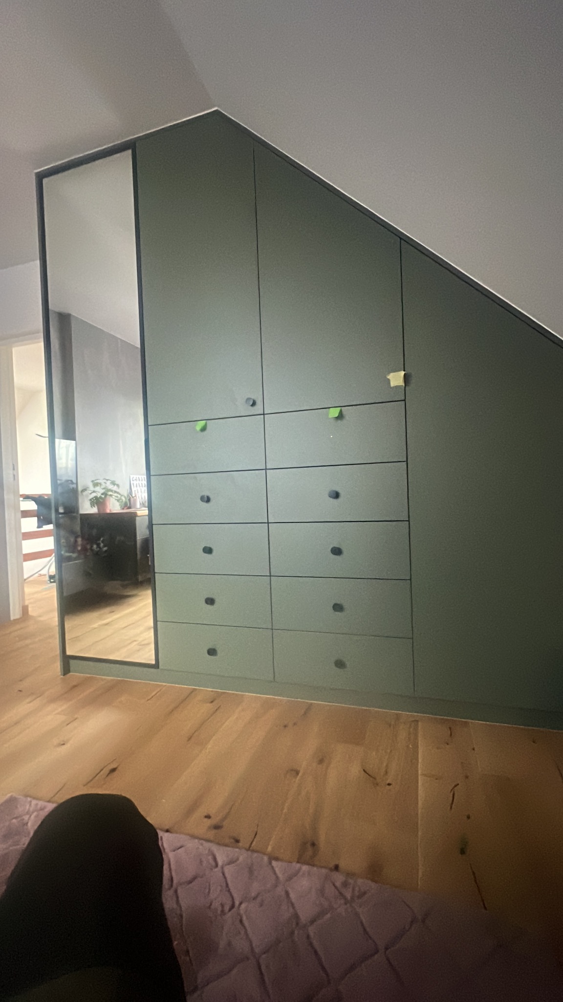 Zabudowa szafy wnękowej w kolorze oliwkowym z lustrem na drzwiach, dopasowana do skosu dachu, z widocznymi drobnymi karteczkami samoprzylepnymi na frontach.