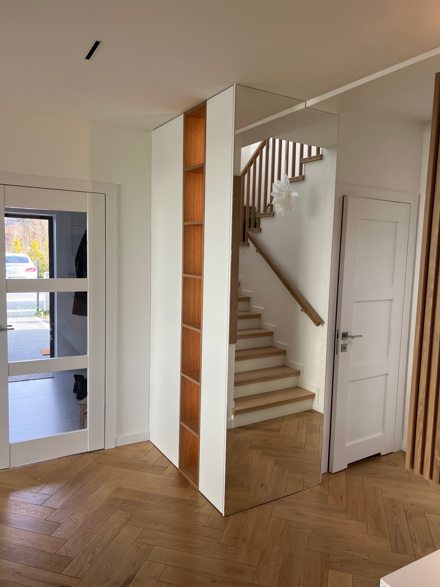 Nowoczesny przedpokój z lustrzaną szafą narożną i drewnianymi półkami, parkiet w jodełkę, widok na schody i drzwi wejściowe.