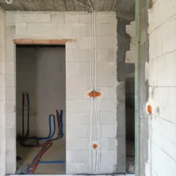 Surowy stan: ściany z betonu komórkowego z widocznymi przewodami elektrycznymi w białych peszlach, puszkami instalacyjnymi w kolorze pomarańczowym, oraz rurami instalacji wodnej w kolorach...