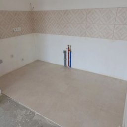 PIO-TREX WYKONCZENIA WNĘTRZ - Przygotowana do dalszych prac wykończeniowych ściana z ułożonymi płytkami dekoracyjnymi i podłoga wyłożona jasnymi płytkami w nowym pomieszczeniu, widoczne rury instalacji wodnej i gniazdka...