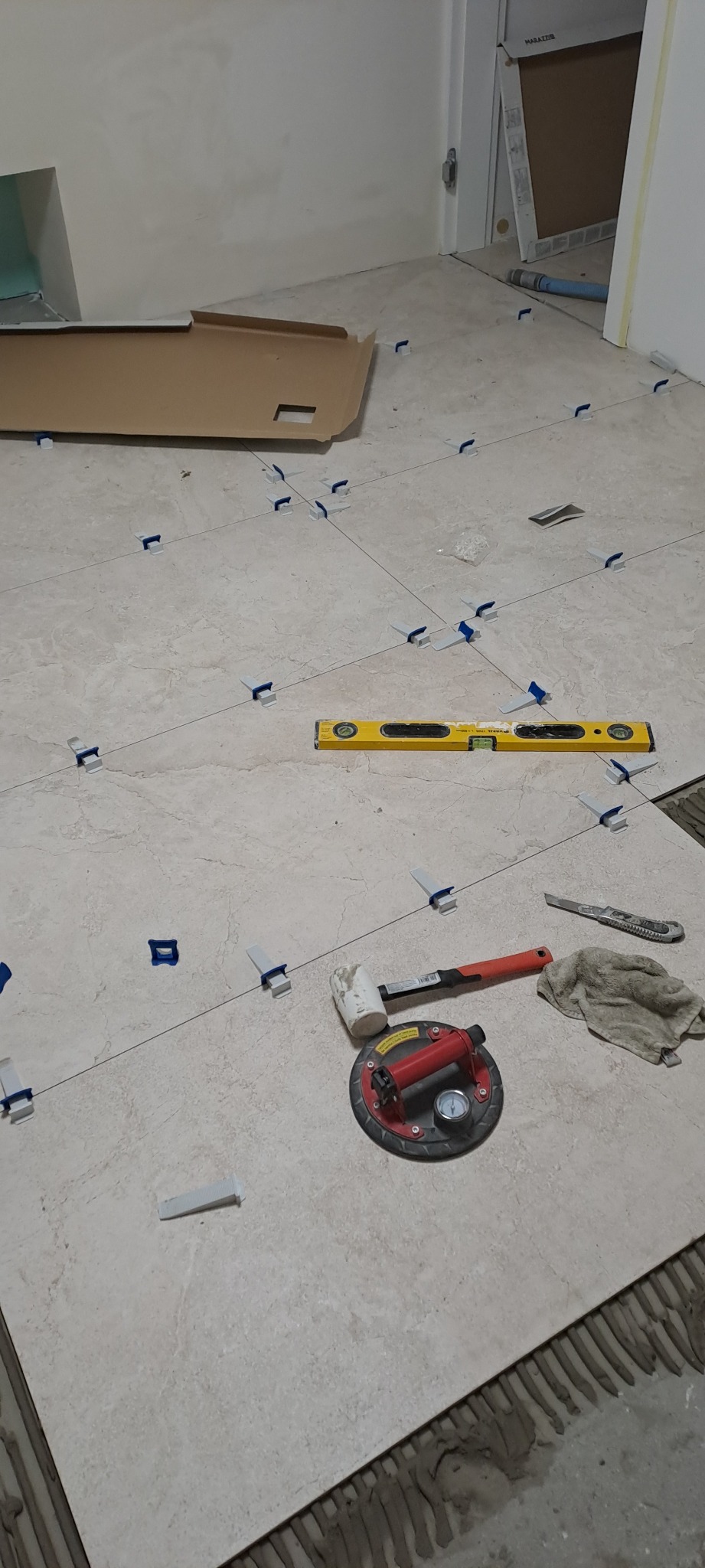 Układanie beżowych płytek podłogowych z użyciem poziomicy, przyssawki, młotka i klipsów poziomujących, widoczna niezagruntowana podłoga i karton po płytkach w tle.