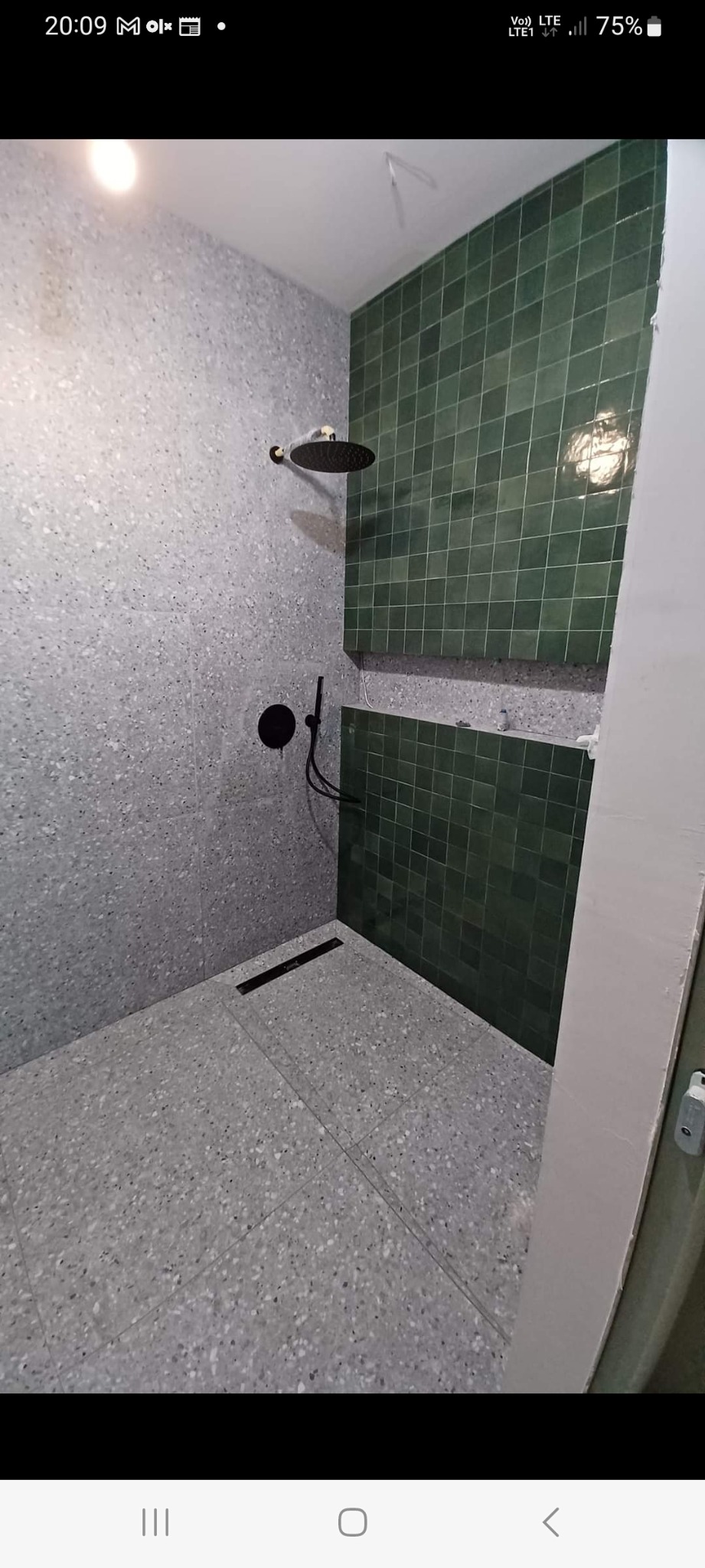 Nowoczesna kabina prysznicowa z szarą, lastrykową posadzką i ścianami oraz fragmentem ściany wyłożonej ciemnozieloną, kwadratową glazurą; czarna deszczownica i bateria podtynkowa.