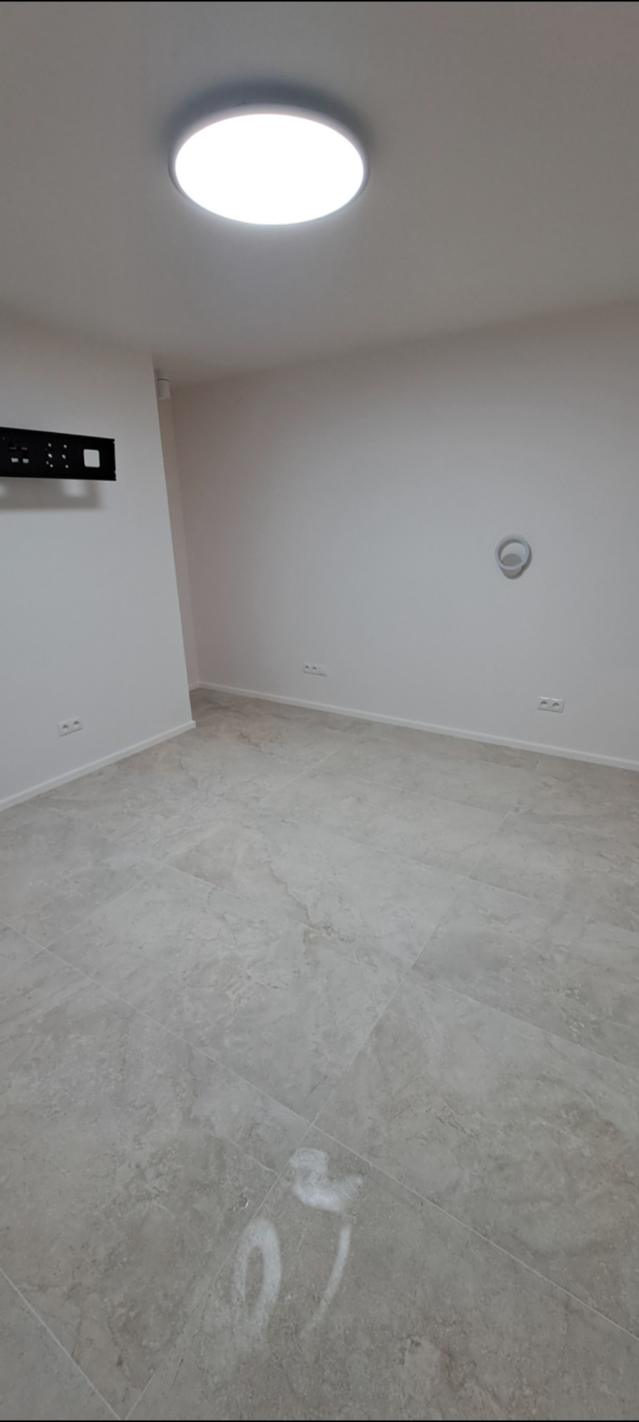 Wykończone wnętrze pokoju z szarymi płytkami podłogowymi imitującymi marmur, białe ściany z gniazdkami elektrycznymi, zamontowany uchwyt na telewizor i okrągła lampa sufitowa.