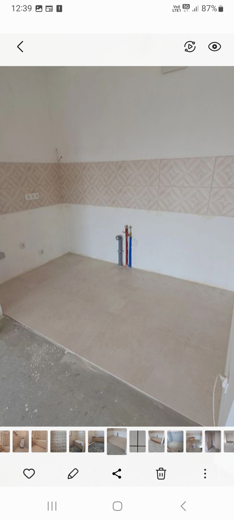 Przygotowana do dalszych prac wykończeniowych ściana z ułożonymi płytkami dekoracyjnymi i podłoga wyłożona jasnymi płytkami w nowym pomieszczeniu, widoczne rury instalacji wodnej i gniazdka...