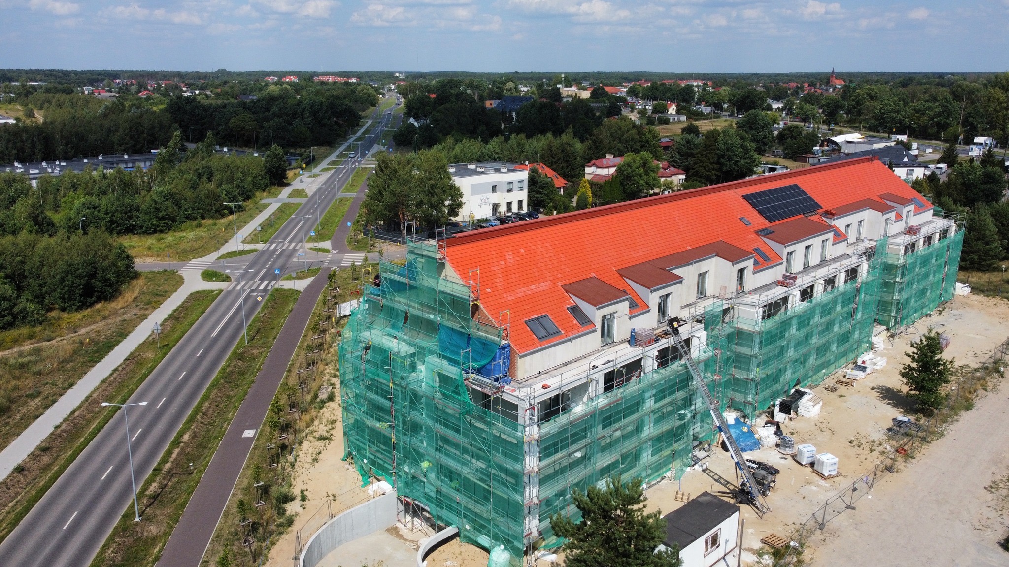 Wielopiętrowy budynek mieszkalny w trakcie prac elewacyjnych, otoczony rusztowaniami i siatką ochronną, z widocznym nowym, czerwonym dachem z panelami słonecznymi, ujęcie z drona, w tle droga...