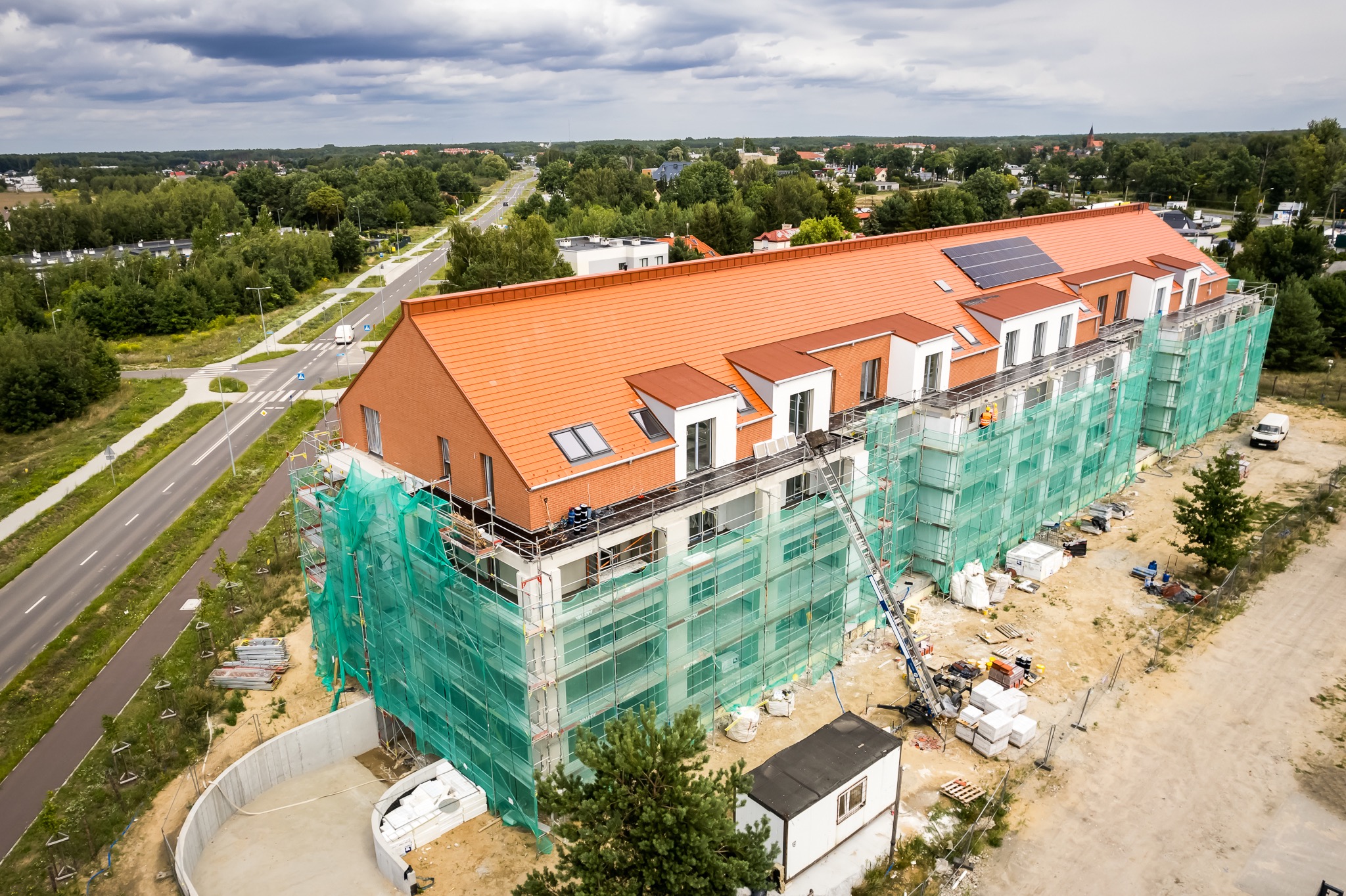 Wysoki budynek w trakcie budowy, z ceglaną elewacją i pomarańczowym dachem, otoczony rusztowaniami zabezpieczonymi zieloną siatką, widok z lotu ptaka na tle zieleni i drogi.