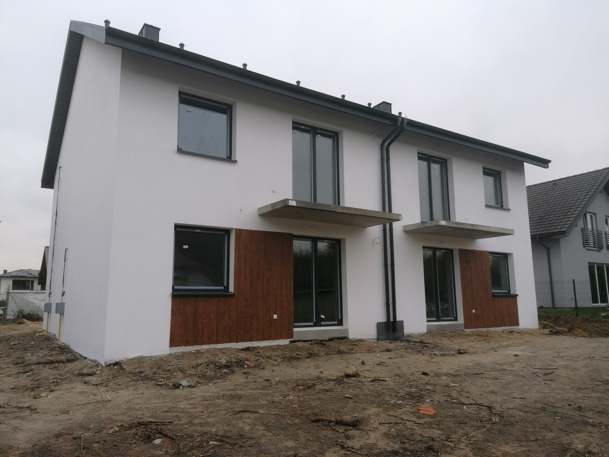 Nowoczesny, dwusegmentowy dom bliźniak z białymi ścianami i brązowymi panelami dekoracyjnymi, czarnymi ramami okiennymi i dachem, w trakcie prac wykończeniowych na zewnątrz.
