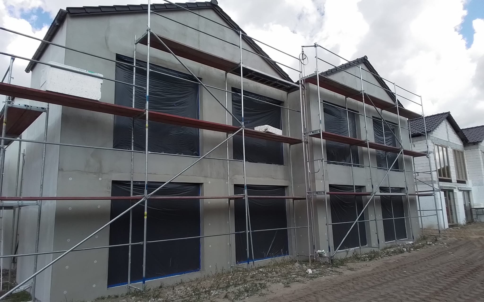 Szare, dwukondygnacyjne budynki w trakcie prac elewacyjnych, osłonięte rusztowaniami i czarną folią okienną, z widocznymi fragmentami ocieplenia i świeżego tynku.