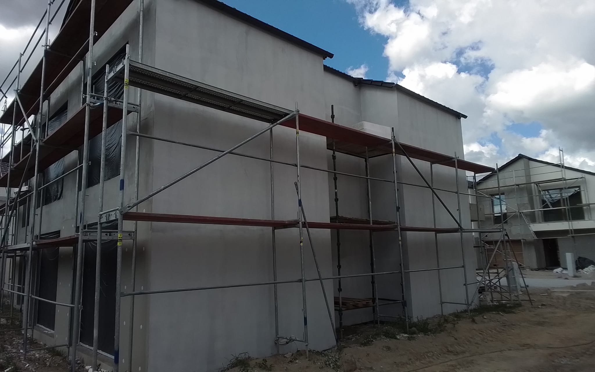Budynek w trakcie tynkowania elewacji, widoczne rusztowania, okna zabezpieczone folią, świeży tynk w kolorze szarym, w tle błękitne niebo z chmurami.