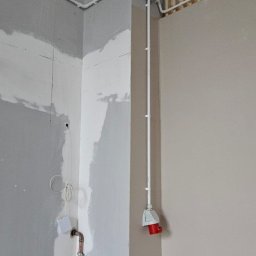 kW Usługi Elektryczne Wojciech Puszkiewicz - Białe rury elektryczne prowadzone po ścianie w rogu pomieszczenia w trakcie wykańczania. Widoczna puszka elektryczna z czerwonym gniazdem.