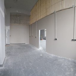 kW Usługi Elektryczne Wojciech Puszkiewicz - Wnętrze w trakcie remontu: widoczne kable elektryczne prowadzone po ścianach, puszki instalacyjne i drewniane elementy dekoracyjne pod sufitem. Szare płytki na podłodze.
