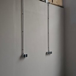 kW Usługi Elektryczne Wojciech Puszkiewicz - Szare przewody elektryczne w metalowych rurach prowadzone po ścianie w nowym budynku, z widocznymi puszkami i gniazdami. Ściana pomalowana na szaro.