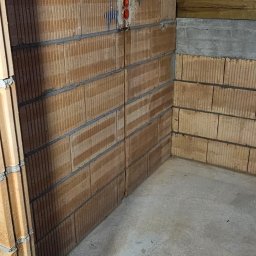 kW Usługi Elektryczne Wojciech Puszkiewicz - Wewnętrzny róg surowego pomieszczenia z instalacją elektryczną w ścianie z cegły dziurawki. Widoczne przewody i gniazdko w kolorze czerwonym.