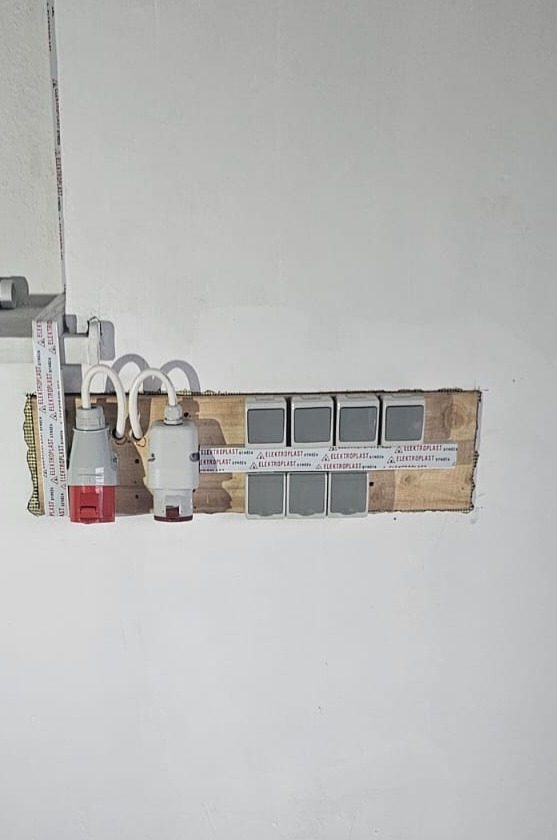 Nierówna ściana w trakcie prac elektrycznych, widoczne gniazda siłowe z wtyczkami, przełączniki światła zamontowane na drewnianej listwie, zabezpieczone taśmą ostrzegawczą z logo producenta.