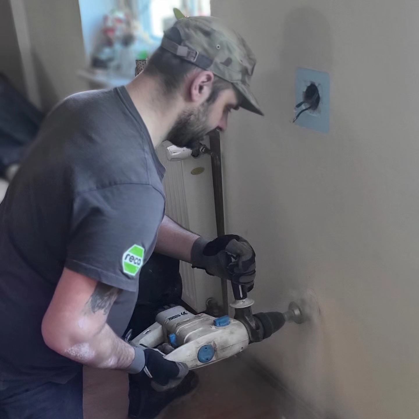 Elektryk w czapce moro wierci otwór w ścianie w pobliżu istniejącego gniazdka elektrycznego, używając wiertarki udarowej z nasadką do wycinania otworów.