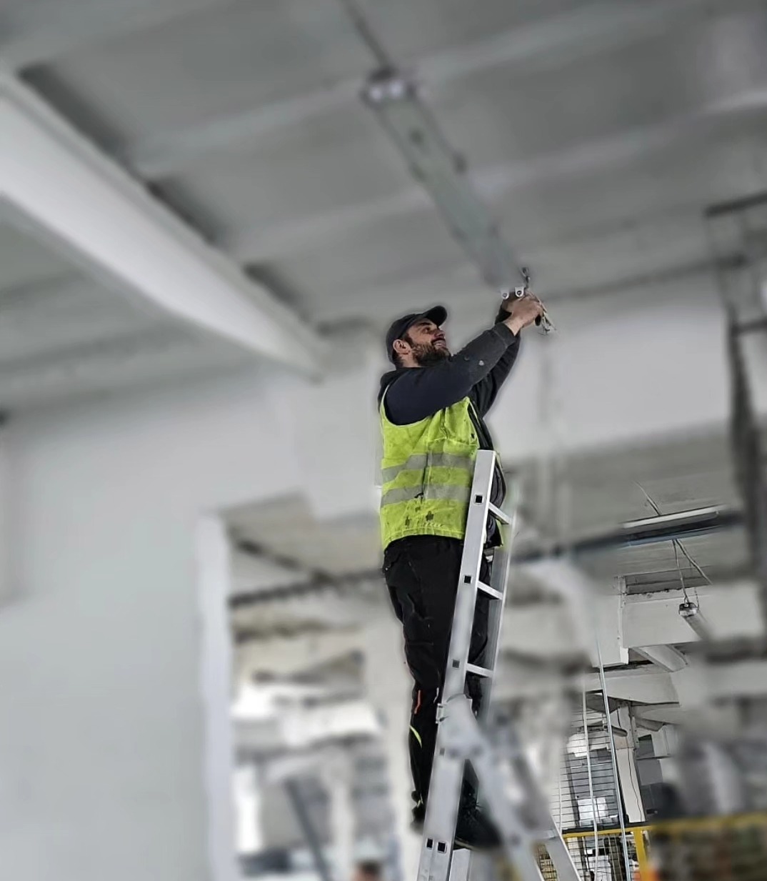 Elektryk w kamizelce odblaskowej na aluminiowej drabinie montuje oświetlenie w hali, widoczne elementy konstrukcji sufitu.