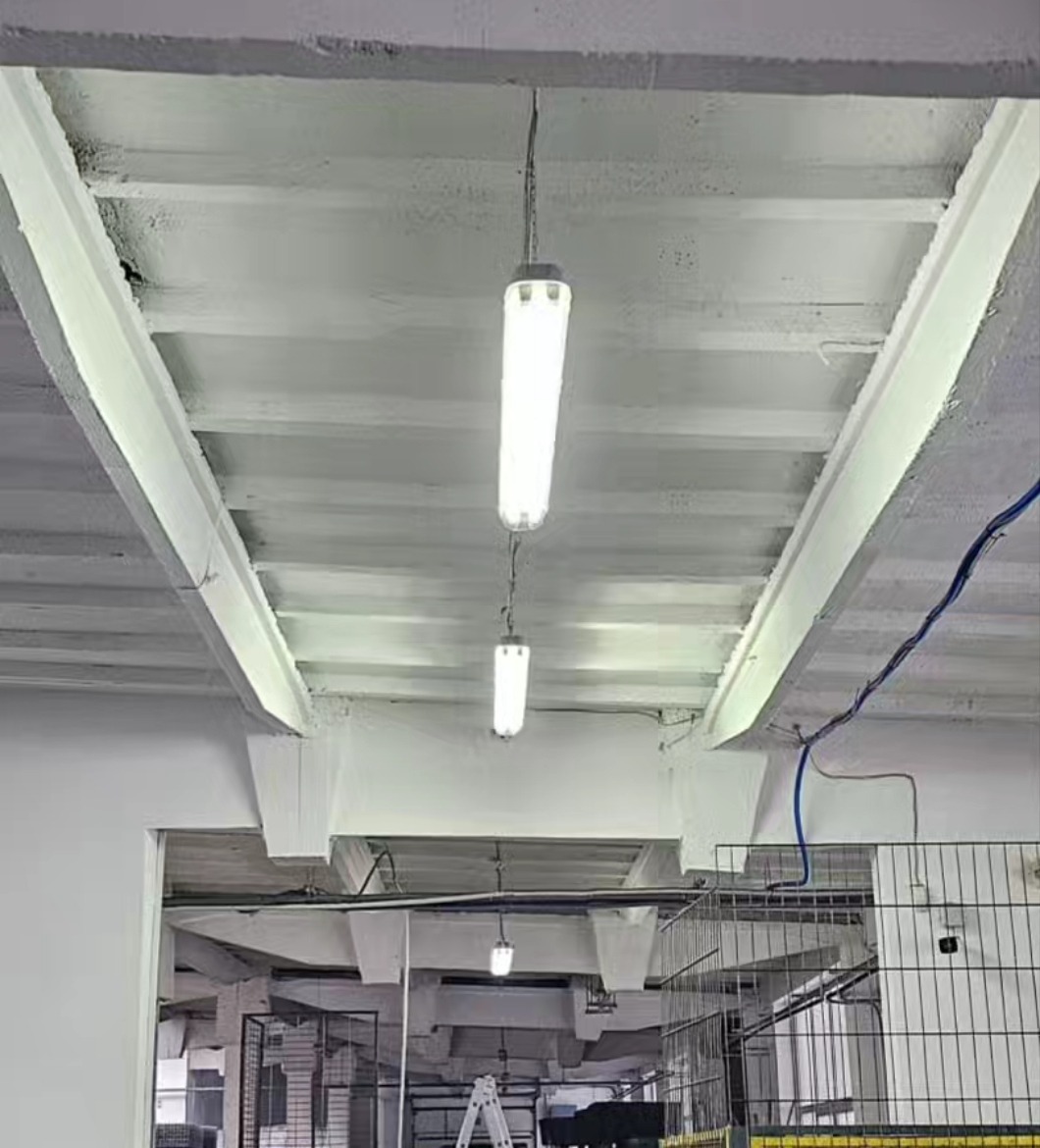 Równomiernie rozmieszczone, podwieszone lampy fluorescencyjne o cylindrycznym kształcie, oświetlające halę produkcyjną w Różańsku, widoczne kable i konstrukcja sufitu.