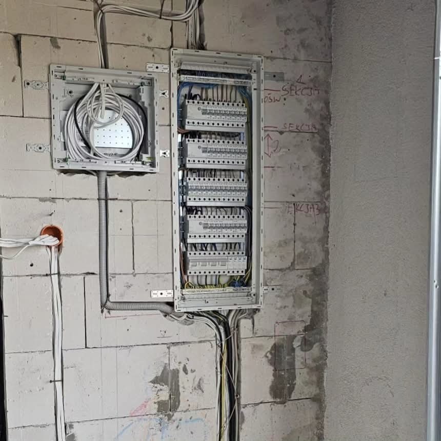 Rozdzielnica elektryczna z okablowaniem na surowej ścianie z betonu komórkowego. Widoczne kable i przewody w peszlach. Stan surowy, instalacja w trakcie montażu.