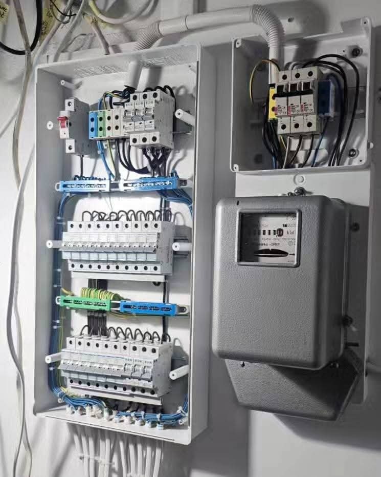 Otwarta skrzynka elektryczna z bezpiecznikami, kable w peszlu, oraz licznik energii elektrycznej. Widok z bliska na instalację elektryczną.