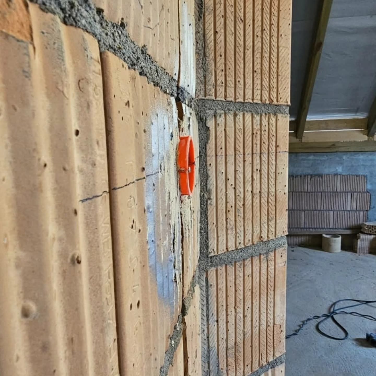 Ściana z pustaków ceramicznych z zamontowaną pomarańczową puszką elektryczną podtynkową, widoczne zaprawy murarskie i przewody w tle.