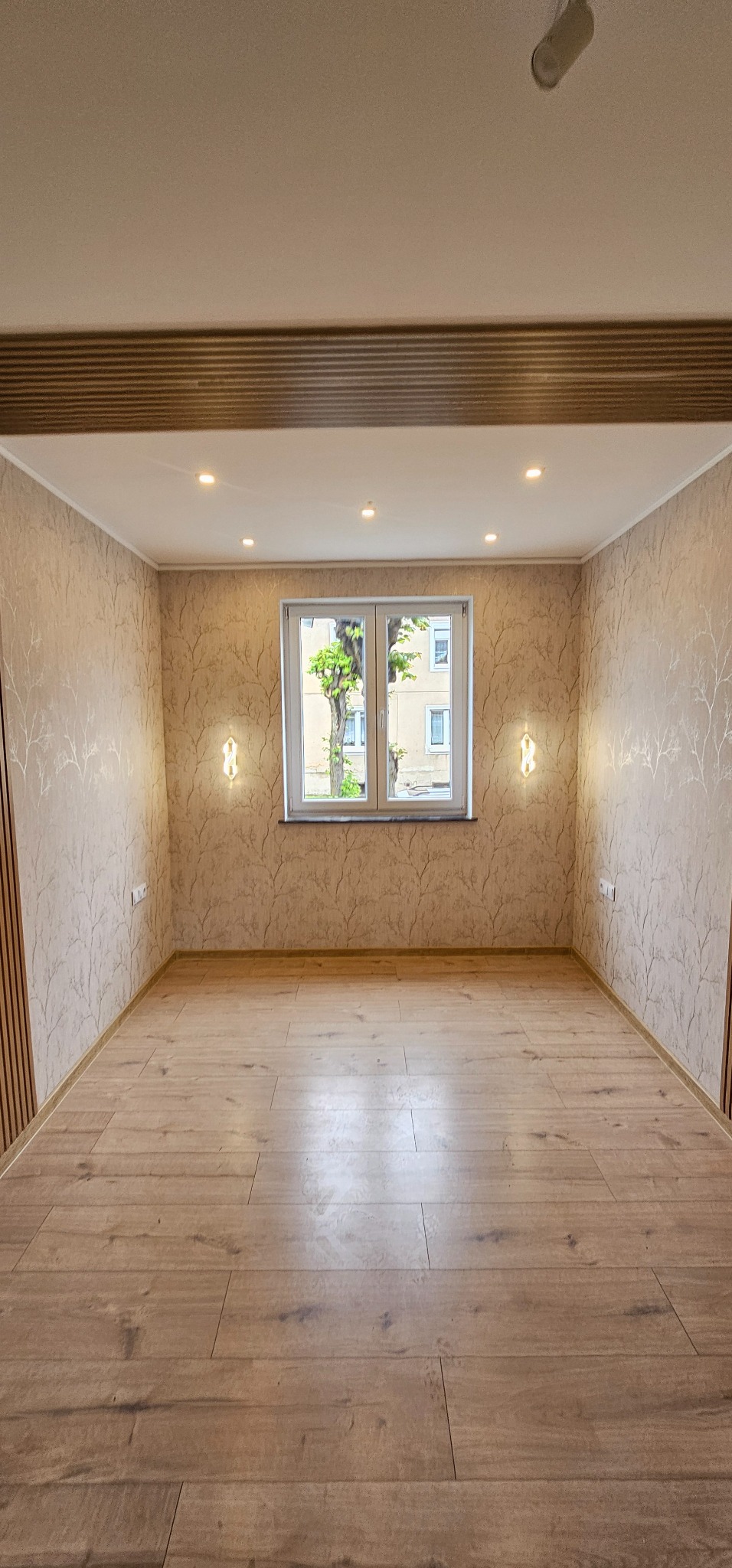 Pusty pokój po remoncie z drewnianą podłogą, wzorzystą tapetą i nowoczesnymi kinkietami po bokach okna. Widok na zewnątrz przez okno.