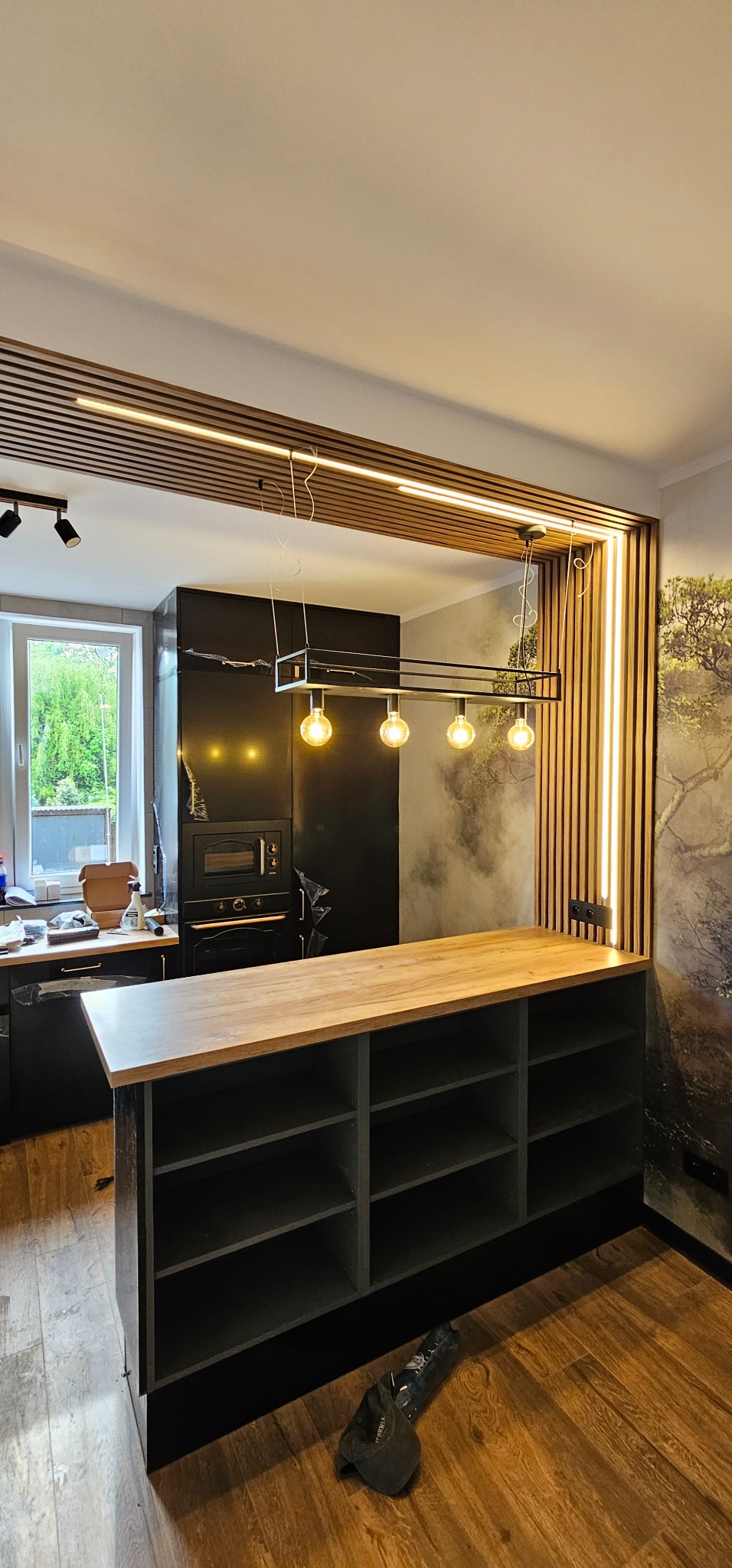 Nowoczesna kuchnia z wyspą, drewnianymi lamelami i oświetleniem LED. Czarny sprzęt AGD i tapeta z motywem drzewa w tle. Lampa wisząca z czterema żarówkami.