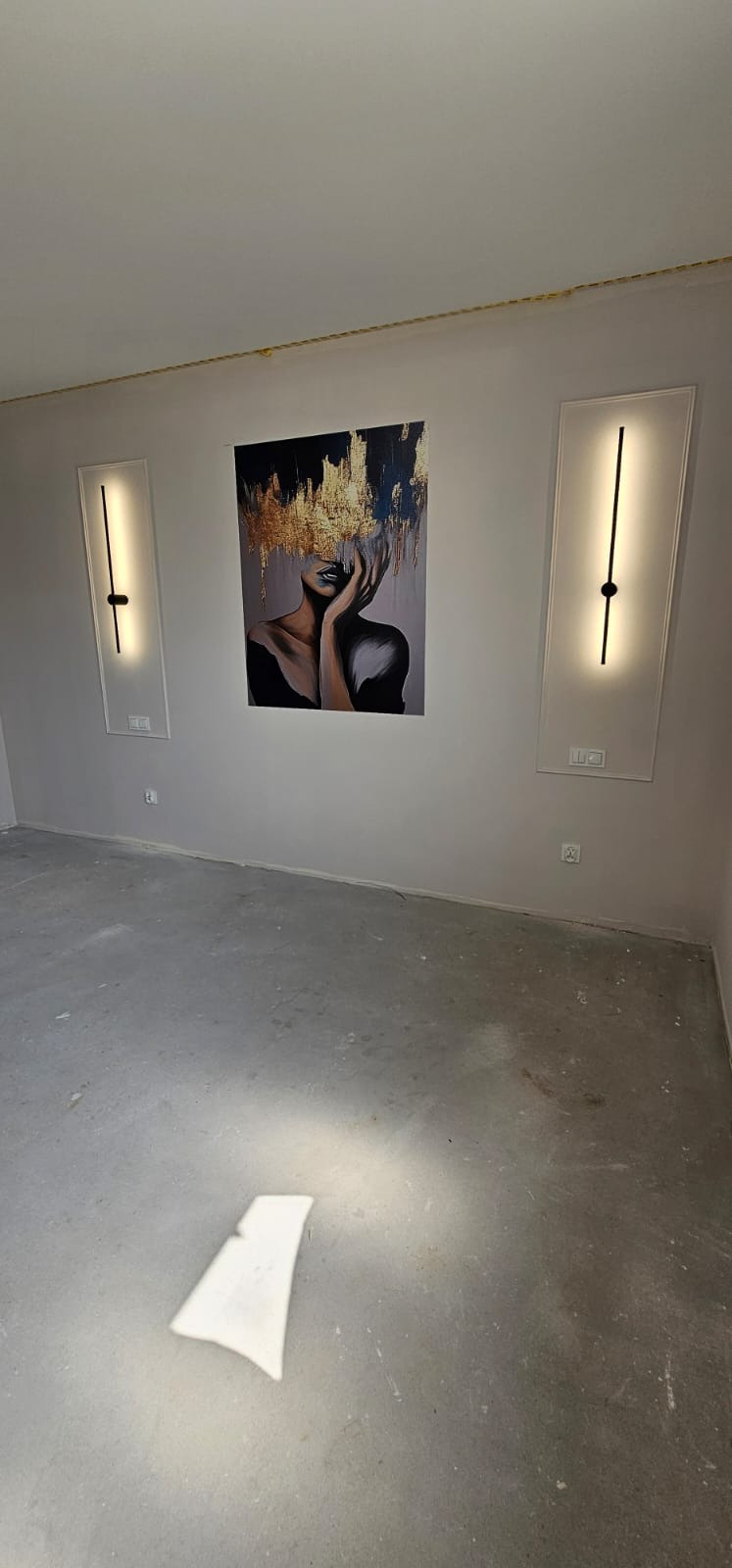 Nowoczesny pokój z dwoma podłużnymi lampami ściennymi po bokach obrazu kobiety z abstrakcyjnym złotym nakryciem głowy. Szare ściany i surowa podłoga.