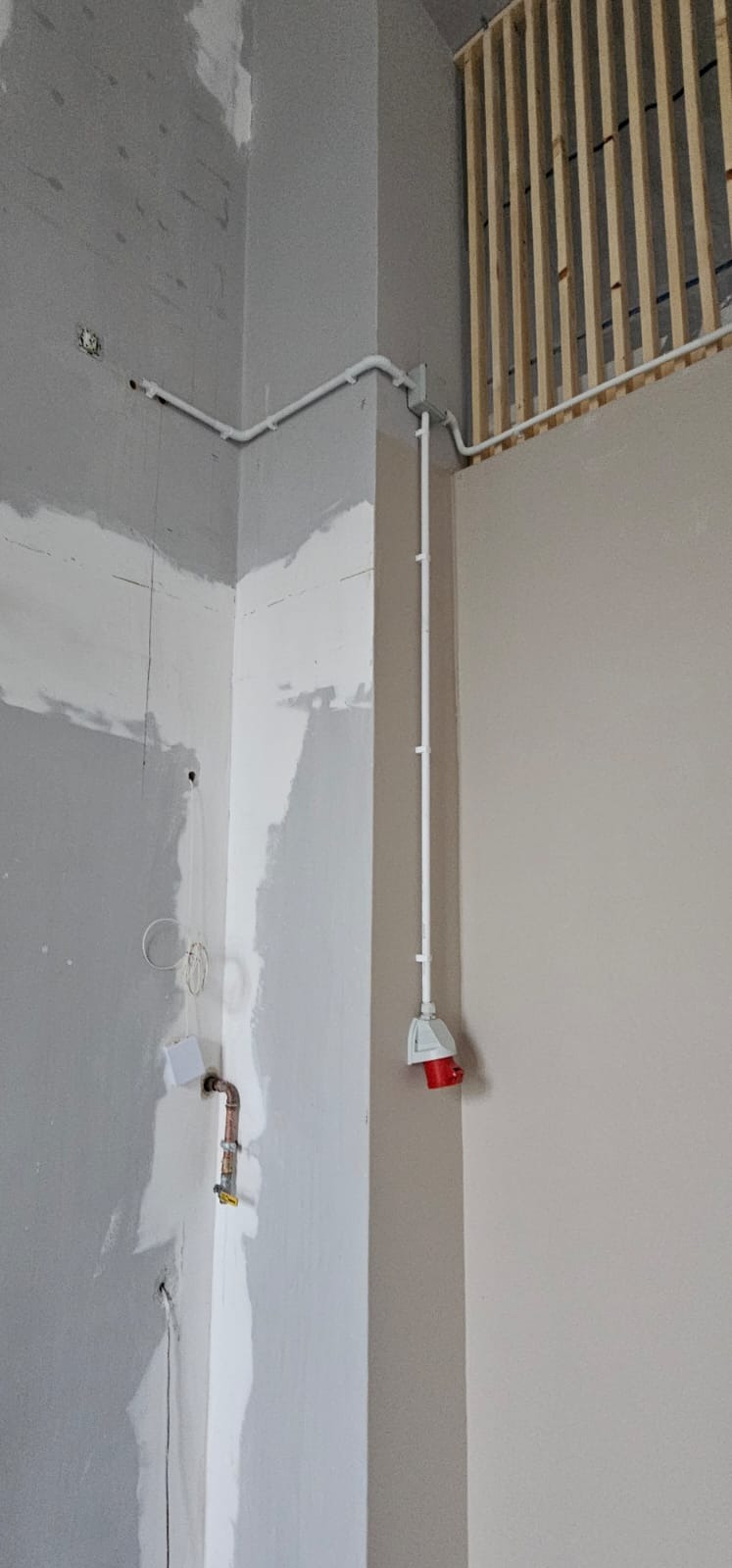Białe rury elektryczne prowadzone po ścianie w rogu pomieszczenia w trakcie wykańczania. Widoczna puszka elektryczna z czerwonym gniazdem.