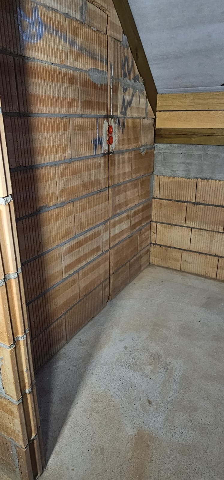 Wewnętrzny róg surowego pomieszczenia z instalacją elektryczną w ścianie z cegły dziurawki. Widoczne przewody i gniazdko w kolorze czerwonym.