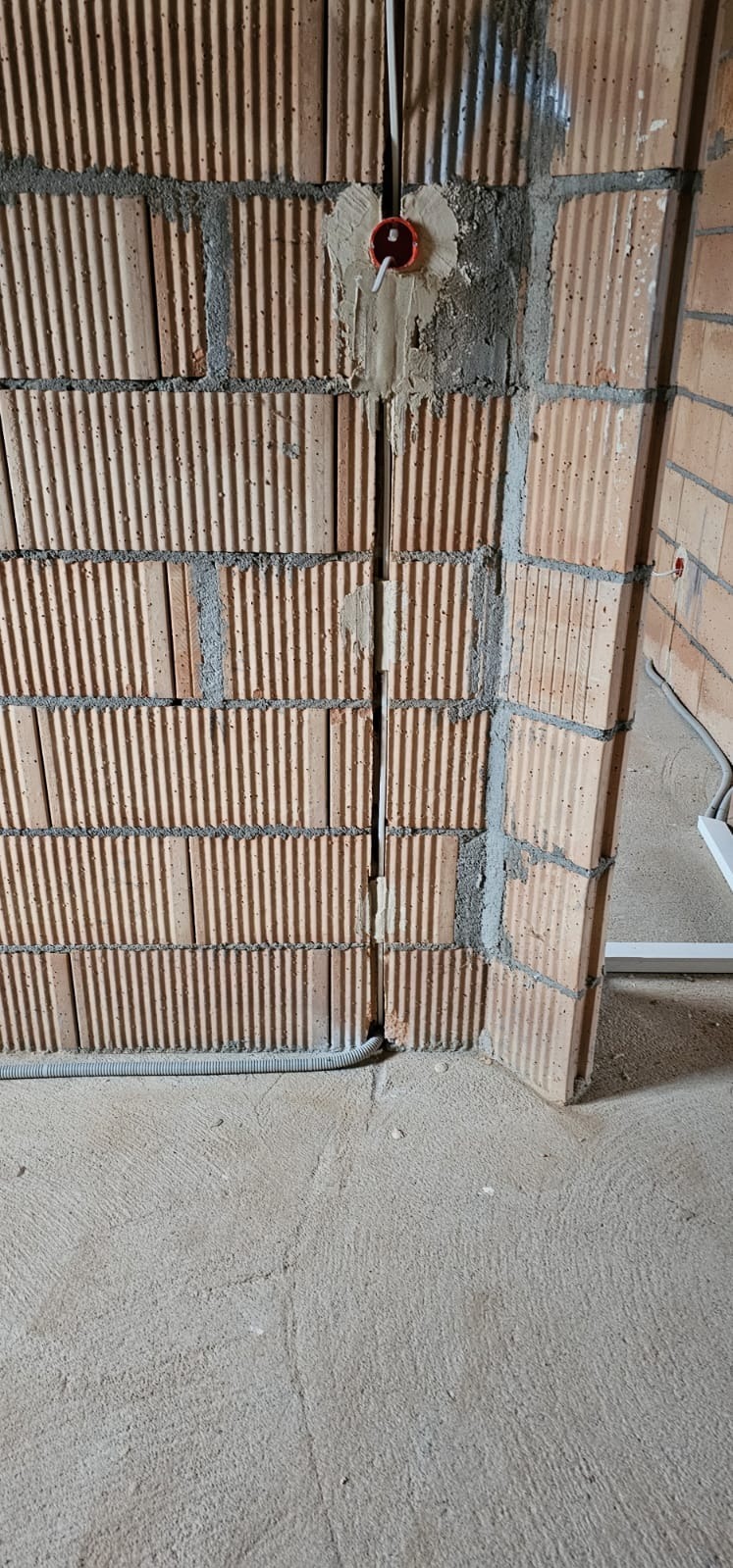 Czerwona puszka elektryczna zamontowana w ścianie z cegły dziurawki, przewody w peszlu ułożone wzdłuż ściany, etap prac instalacyjnych.